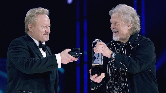 Un homme tient une petite caméra devant le trophée exposé par un autre homme sur une scène, lors d'une soirée de gala.