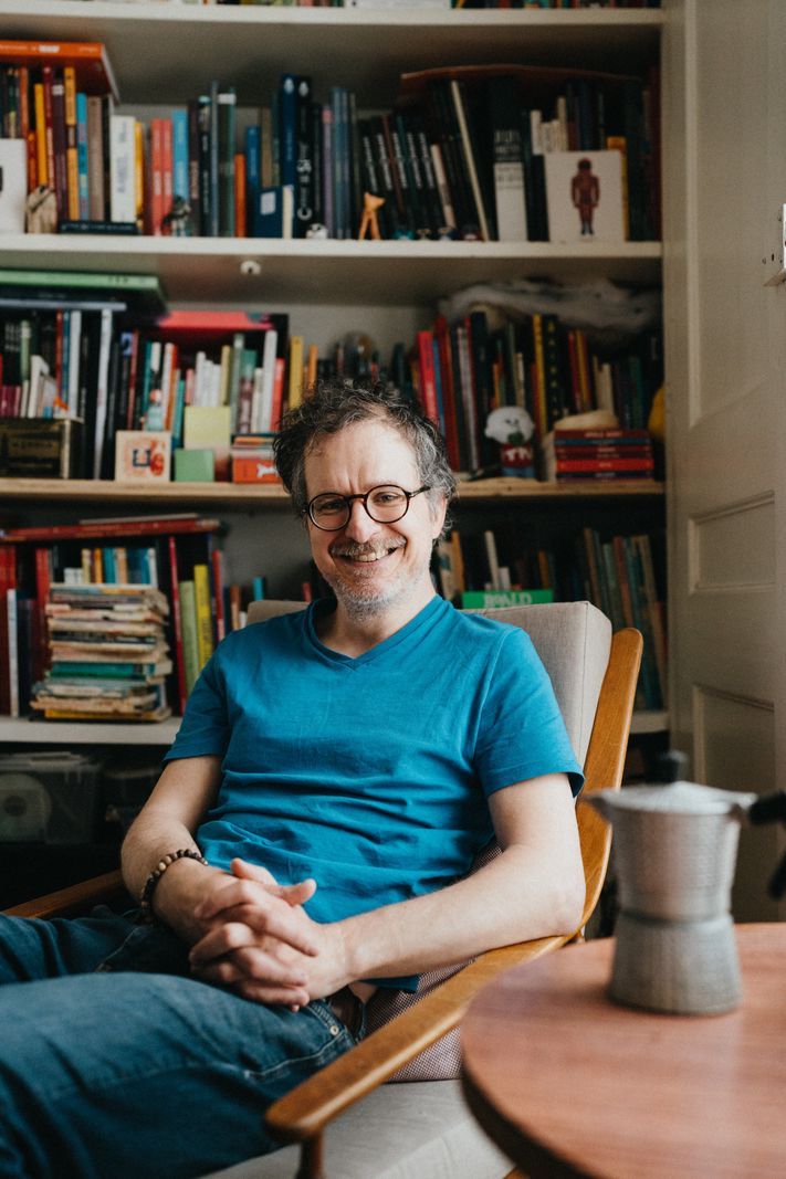 Un homme souriant portant un t-shirt bleu assis sur une chaise devant une bibliothèque bien approvisionnée. Un homme souriant portant un t-shirt bleu assis sur une chaise devant une bibliothèque bien approvisionnée.