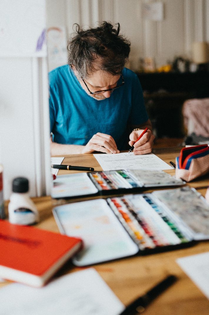 Homme concentré, vêtu d'un t-shirt bleu, dessinant sur un bureau encombré d'aquarelles et de fournitures artistiques. Homme concentré, vêtu d'un t-shirt bleu, dessinant sur un bureau encombré d'aquarelles et de fournitures artistiques.