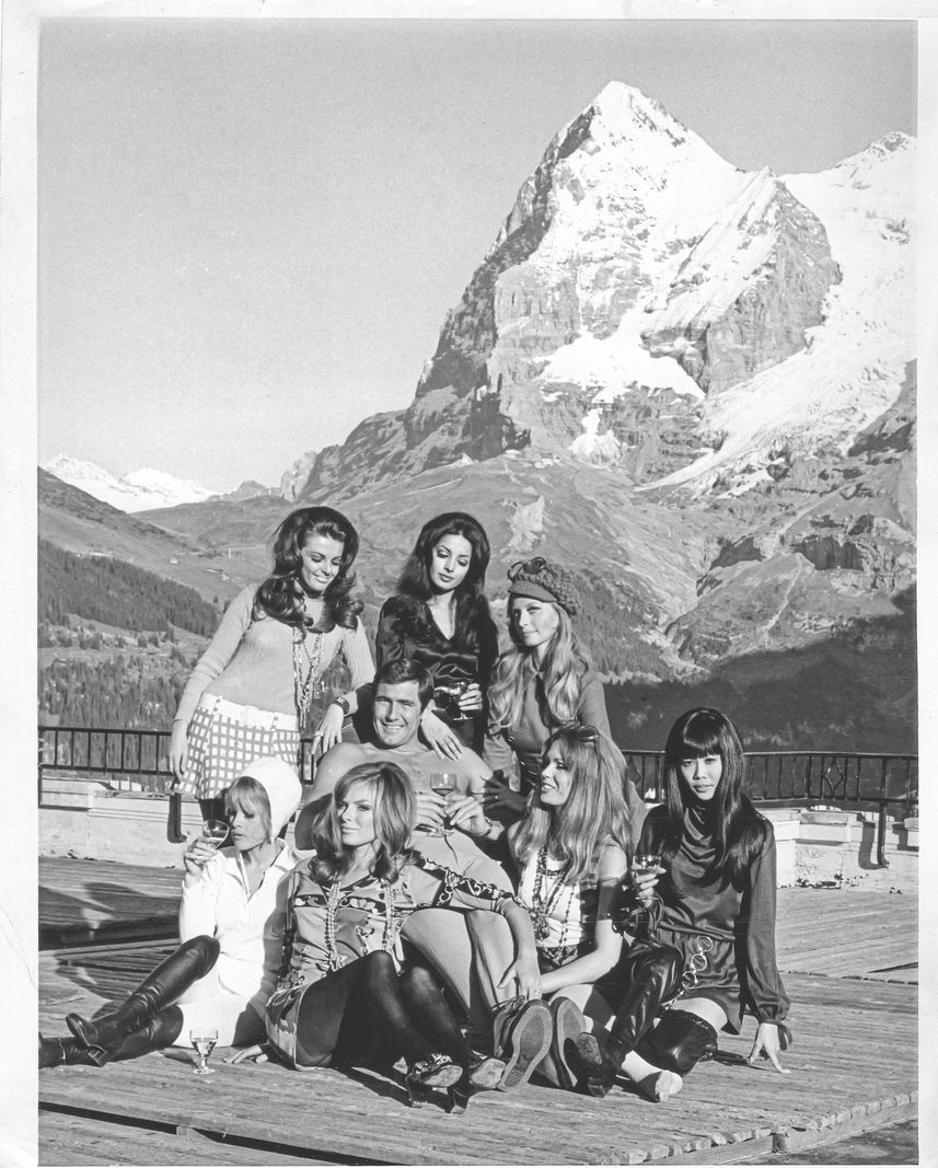 Lazenby entouré de sept filles de Bond devant l'hôtel Palace à Mürren, avec une vue sur le Mont Eiger en arrière-plan. Lazenby entouré de sept filles de Bond devant l'hôtel Palace à Mürren, avec une vue sur le Mont Eiger en arrière-plan.