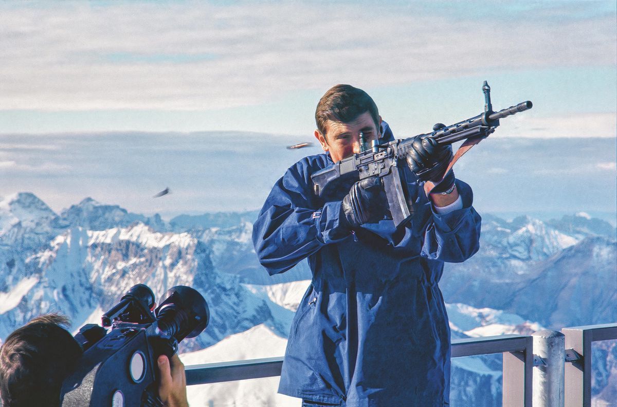 George Lazenby comme James Bond sur le Gloria Piz, tenant un fusil lors d'un tournage, avec les Alpes suisses en arrière-plan. George Lazenby comme James Bond sur le Gloria Piz, tenant un fusil lors d'un tournage, avec les Alpes suisses en arrière-plan.