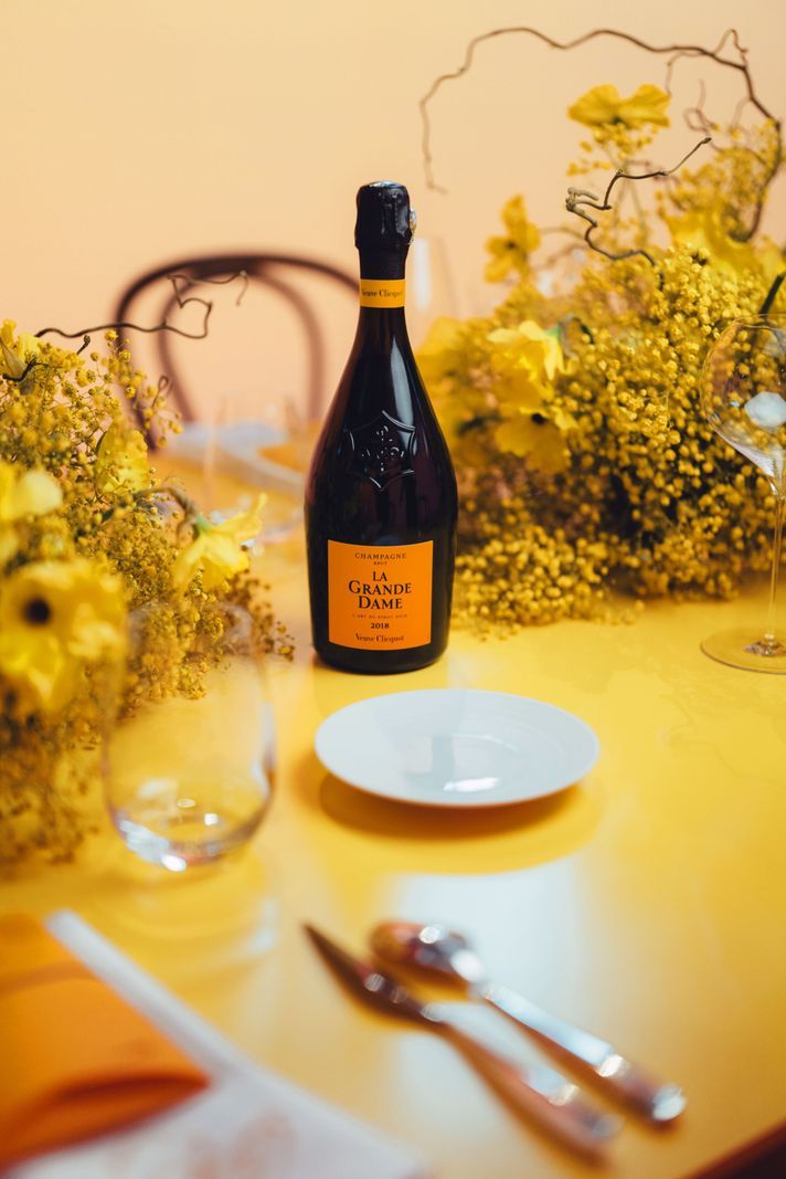 Bouteille de champagne La Grande Dame sur une table décorée de fleurs jaunes, de verres et de couverts. Bouteille de champagne La Grande Dame sur une table décorée de fleurs jaunes, de verres et de couverts.