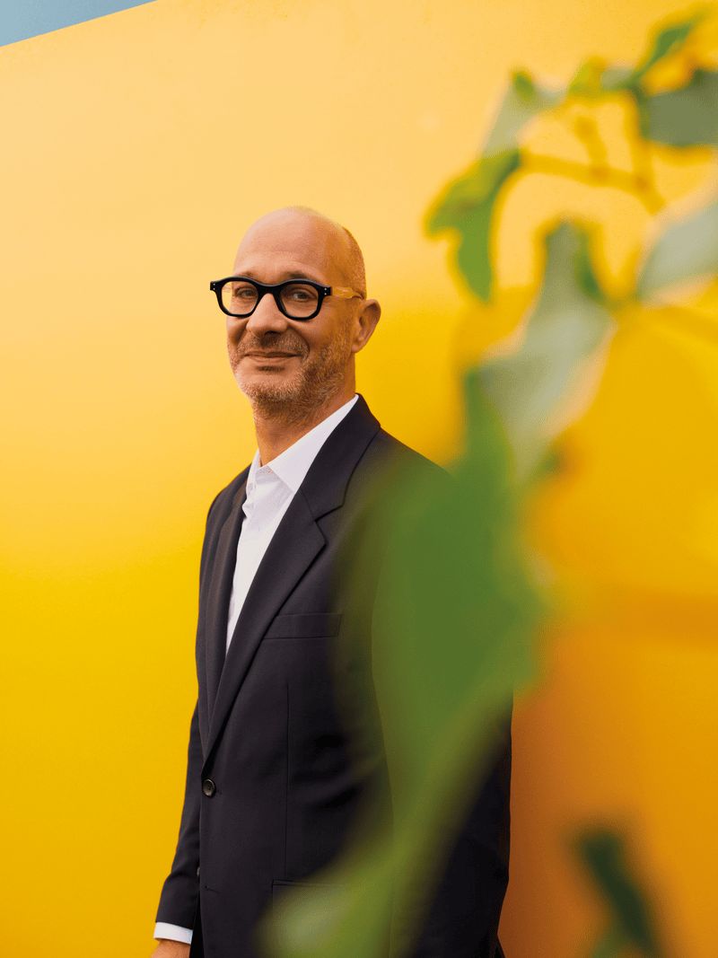 Homme aux lunettes noires, vêtu d'un costume sombre, debout devant un mur jaune vif avec une plante floue au premier plan. Homme aux lunettes noires, vêtu d'un costume sombre, debout devant un mur jaune vif avec une plante floue au premier plan.